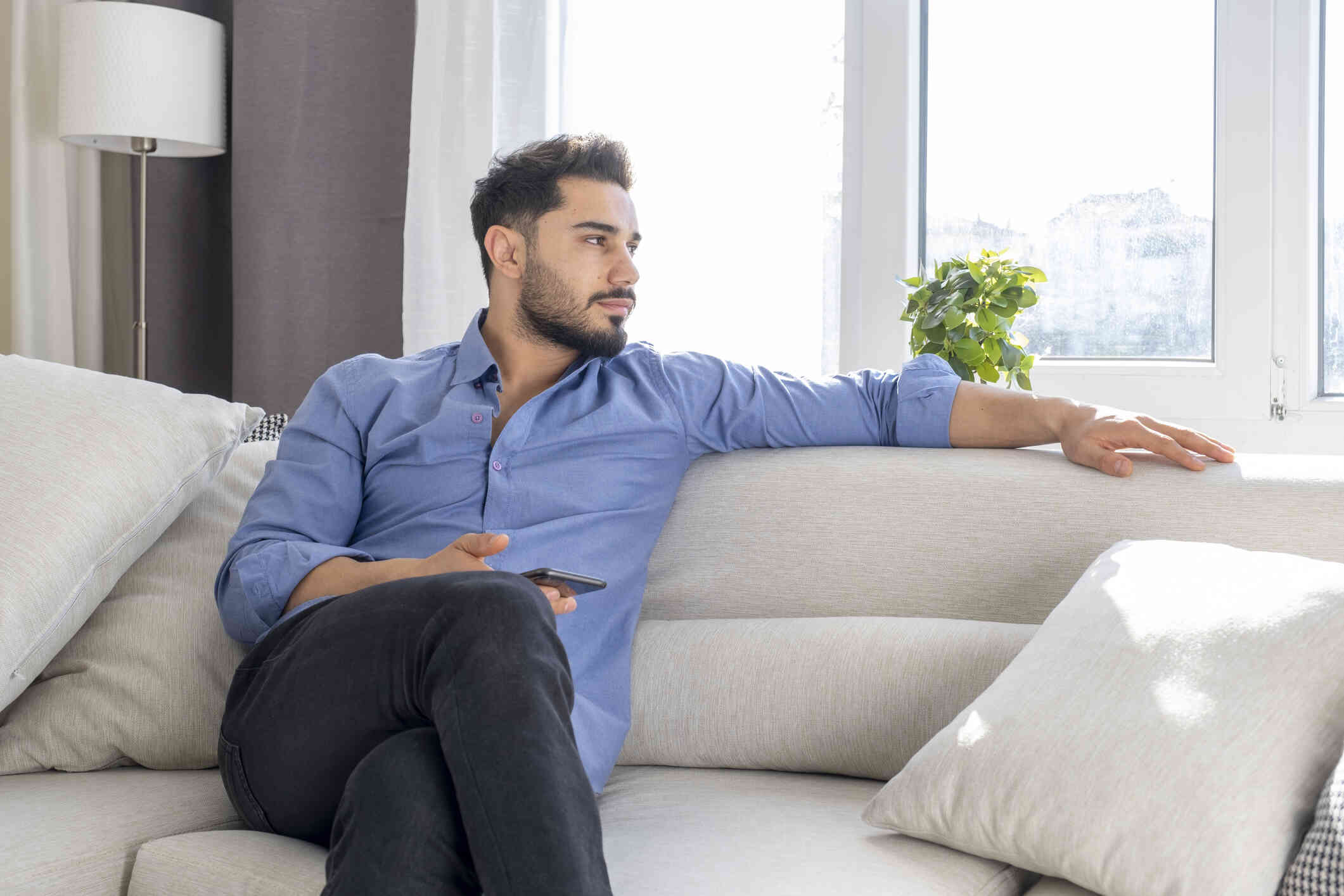 A man ina blue button down shirt sits on the couch with his cellphone in his hand while gazing off deep in thought.